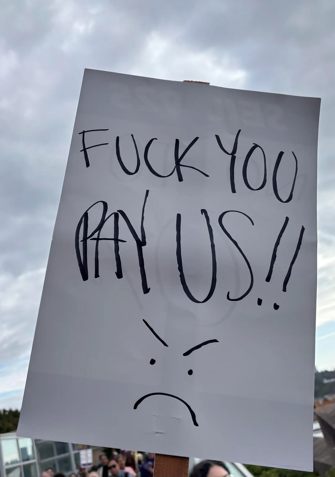 Handwritten protest sign reading “Fuck you, pay us!!” with an angry face drawn below.