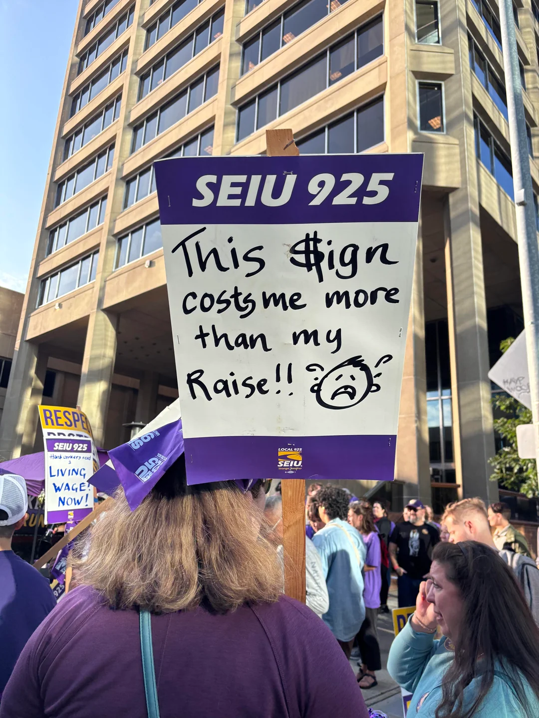 SEIU 925 protest sign reading “This sign costs me more than my raise!!” held above a crowd outside a tall building.