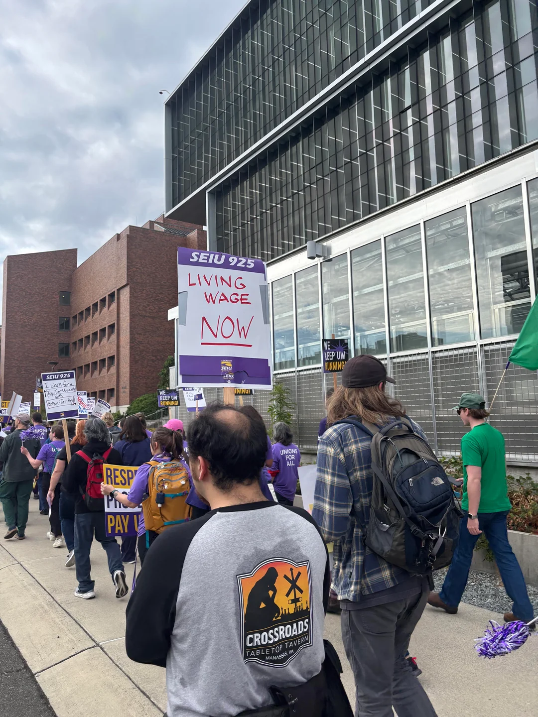 Marchers carry SEIU 925 signs, including “Living wage now,” past a glass-fronted campus building.