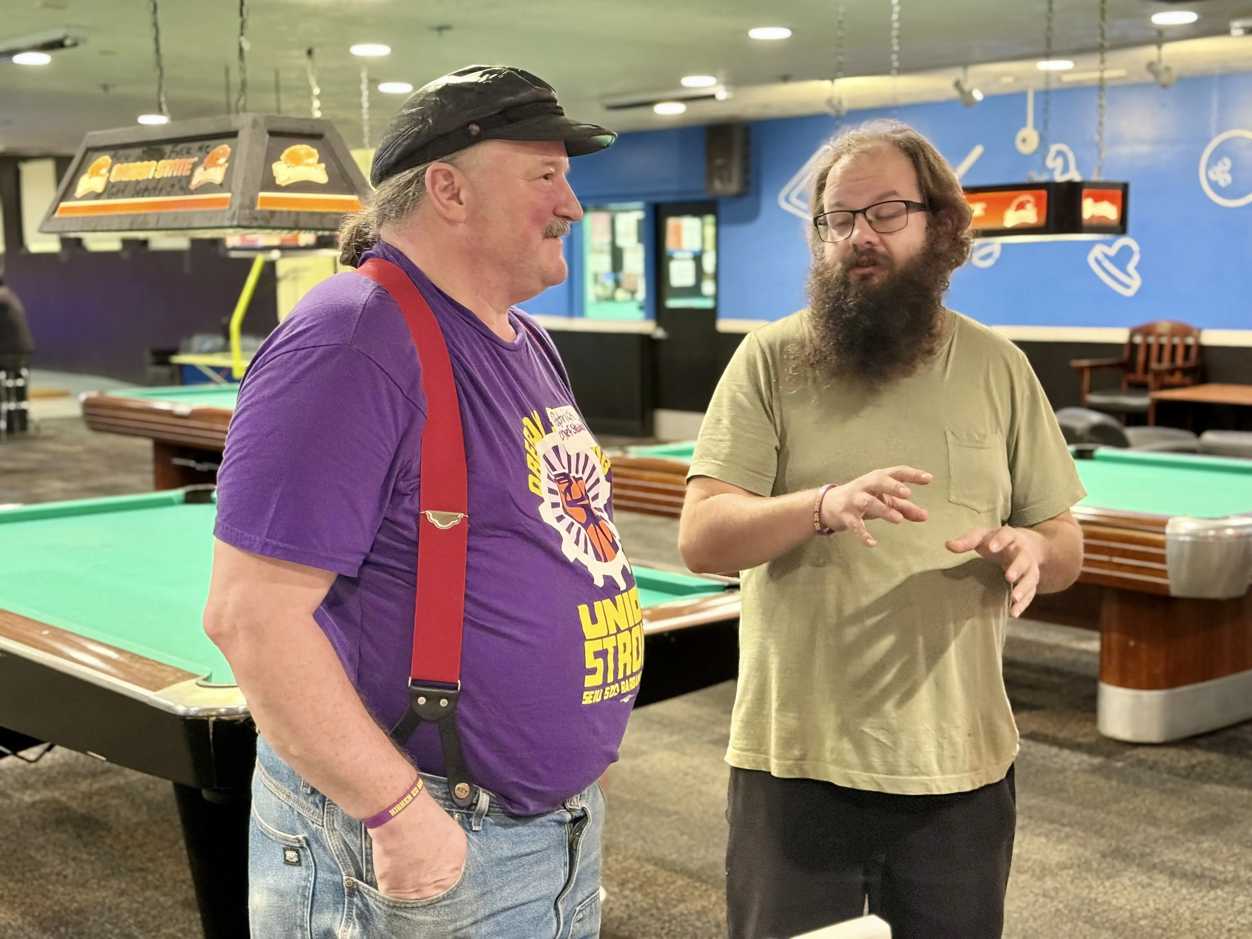 Two men stand next to a green pool table, engaged in conversation. The man on the left wears a purple shirt with red suspenders, and the man on the right has a long beard and is gesturing with his hands.