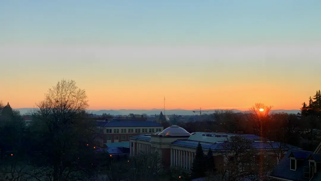 A vibrant sunrise over the Memorial Union building, featuring a gradient orange sky, distant mountains, and silhouette trees on a clear and cold winter morning.