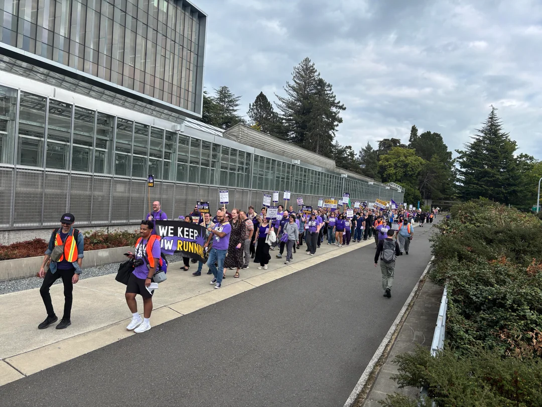 A long procession of marchers in purple follows a “We keep UW running” banner along a campus walkway.
