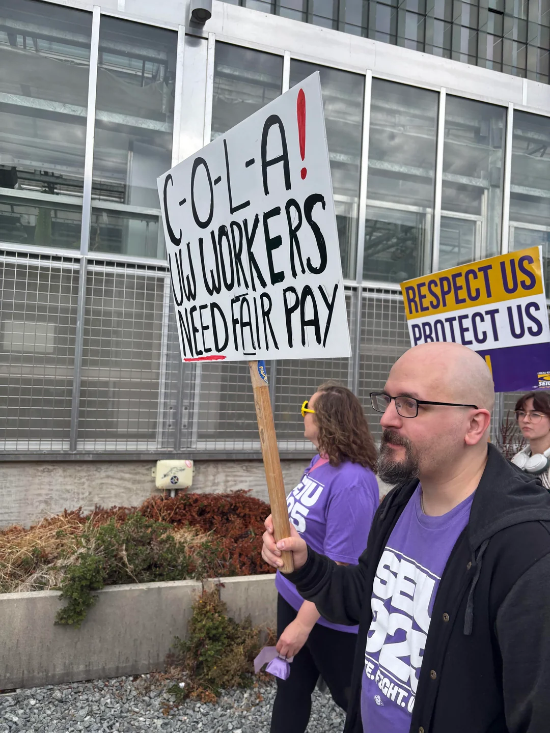 Marcher holds a sign reading “C-O-L-A! UW workers need fair pay,” beside another demanding respect and protection.